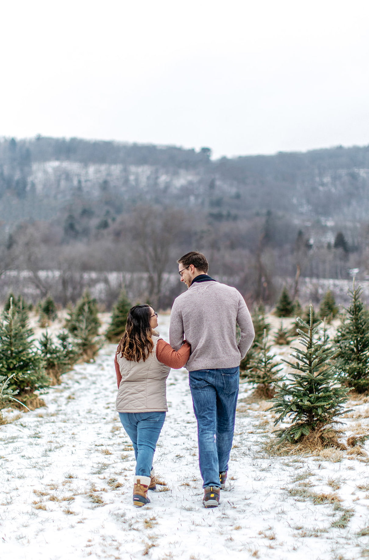 couple holds hands and walks down a path in a snowy field of a christmas tree farm