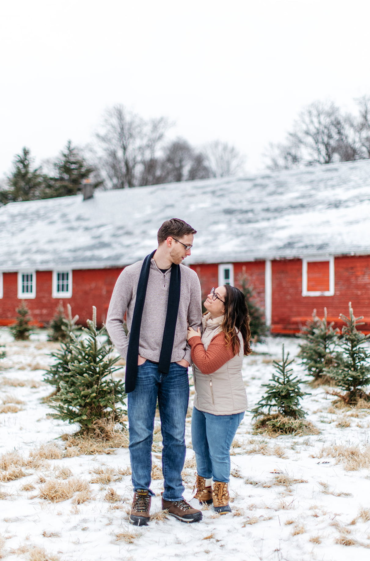 couple stands looking at each other in front of a red barn on a christmas tree farm in upstate ny