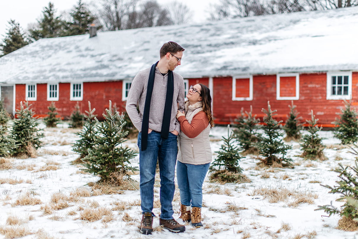 couple stands smiling in front of christmas trees and a red barn for their snowy winter engagement session in upstate ny