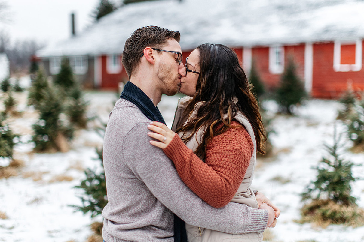 couple kisses standing in front of red barn during snowy winter engagement session on a christmas tree farm in upstate ny